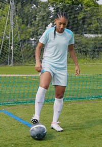 Svetlomodrý športový outfit s krátkymi rukávmi a šortkami, biele podkolienky, čierno-biely futbalový lopta, stojaci na zelenej trávnatej ploche.