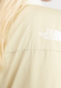 Veste beige avec le logo blanc The North Face sur la poitrine, portée par une personne aux longs cheveux blonds.