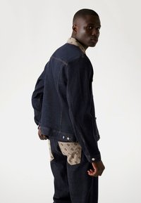 Chaqueta de mezclilla oscura con un cuello y bolsillos de un patrón beige contrastante. Presenta botones y un ajuste estructurado, mostrada de lado.