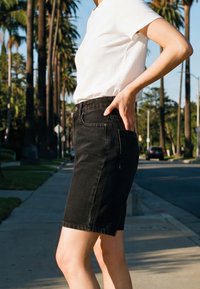 Personne debout sur un trottoir portant un t-shirt blanc et une jupe en jean noire, avec une main posée sur la hanche.