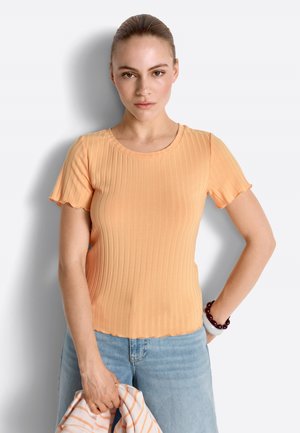 Kobieta z włosami zaczesanymi do tyłu, ubrana w brzoskwiniowy, prążkowany top z krótkim rękawem oraz jasnoniebieskie jeansy, trzymająca w jednej ręce pasiasty element garderoby.