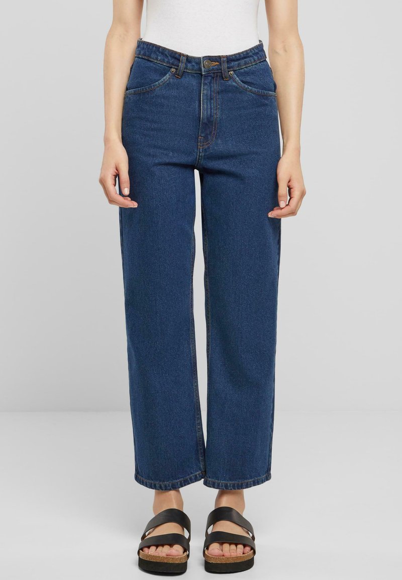 Jean en denim bleu à taille haute et coupe ample avec des coutures visibles, des poches et une fermeture à bouton. Associé à des sandales noires à lanières.