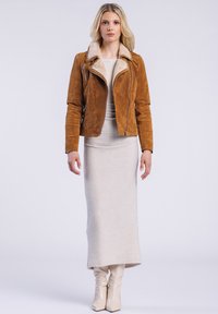 Eine Frau steht selbstbewusst da und trägt eine braune Wildlederjacke über einem beigen Kleid und knielangen, cremefarbenen Stiefeln, vor einem hellen Hintergrund.