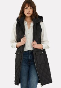 Schwarze gesteppte Weste mit Kapuze, ausgestattet mit einem Reißverschluss und Druckknopftaschen. Getragen über einer weißen Bluse und blauen Jeans, die ein elegantes Design präsentieren.