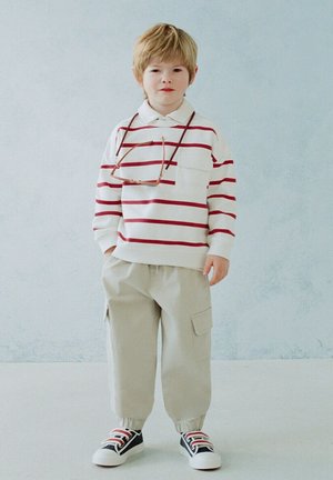 Băiat tânăr în picioare, purtând o bluză albă cu dungi roșii cu mâneci lungi, pantaloni cargo bej, adidași negri și ochelari agățați în jurul gâtului.