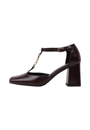 Zapato de tacón alto con tira en T de cuero marrón oscuro, puntera cuadrada y tacón grueso, con un pequeño detalle de cadena dorada.