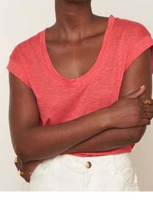 Osoba z założonymi rękami, ubrana w koralową koszulę z krótkim rękawem oraz białe spodnie z detalami w postaci złotych guzików i złotymi pierścieniami na palcach.