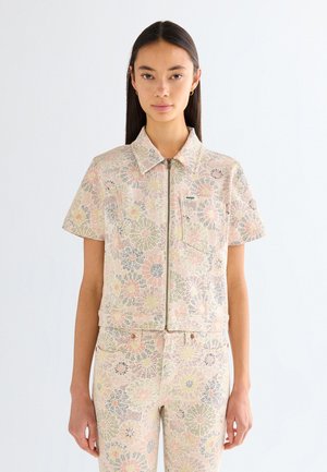 Žena oblečená v krátkym rukávom, na zips kvetinovom saku a zodpovedajúcich nohaviciach v pastelových farbách, stojaca na bielom jednoduchom pozadí.