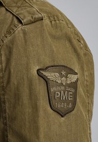 Veste en tissu vert olive avec une épaule matelassée, ornée d'un badge brodé affichant "American Classic PME 1649-A" et un design d'ailes.