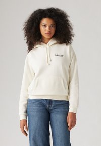 Krämfärgad luvtröja med framficka, dragsko och svart "LEVI'S"-logotyp på bröstet, i kombination med blå vida jeans.