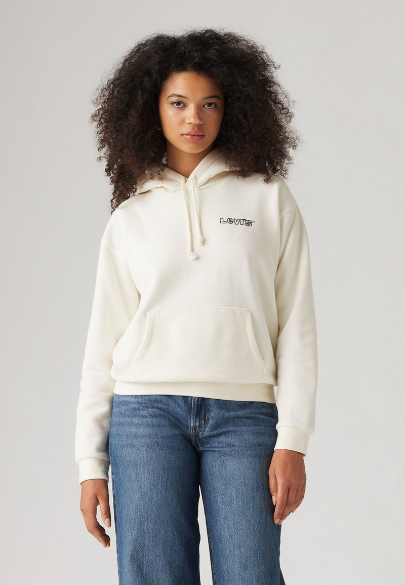 Krämfärgad luvtröja med framficka, dragsko och svart "LEVI'S"-logotyp på bröstet, i kombination med blå vida jeans.