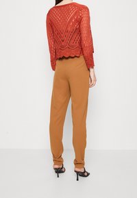 Maglione all'uncinetto in ruggine con orlo a scallop e motivo di lavorazione dettagliato, abbinato a pantaloni marroni a sigaretta e sandali neri con tacco.