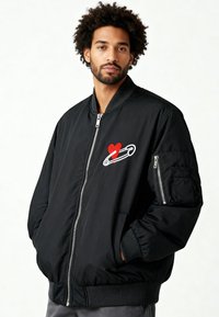 Veste bomber noire avec un motif brodé de cœur rouge et une épingle de sûreté. Fermeture éclair, poches latérales et poignets côtelés. Texture de tissu lisse.