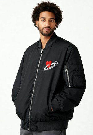 Zwarte bomberjack met een rood hart en veiligheidsspeld geborduurd ontwerp. Ritssluiting, zijzakken en geribbelde manchetten. Soepele stoftextuur.