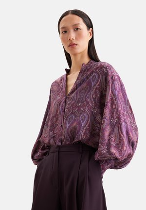 Frau trägt eine locker sitzende lila Paisley-Bluse mit voluminösen Ärmeln und hoch taillierte dunkle Hose, steht vor einem schlichten hellen Hintergrund.