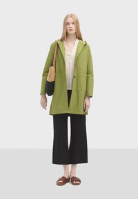 Veste en coton verte avec une capuche, fermeture à bouton et poches avant. Associée à une chemise légère, un pantalon large noir et des sandales marron.
