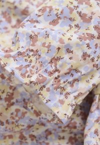 Tissu floral aux couleurs douces et atténuées : jaune pâle, lavande et marron. La texture semble légère avec un détail de volant délicat.