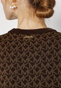 Bruine gebreide trui met een geometrisch patroon in goud, een geribbelde halslijn en een klein gouden logoplaatje aan de achterkant.