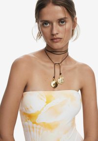 Choker en cuir noir avec deux pendentifs en forme de coquillage dorés. Le premier est lisse, le second présente des détails texturés. Fond blanc.