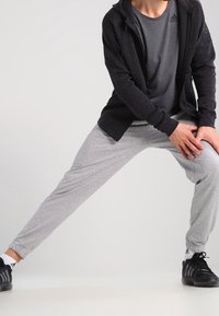 Osoba v šedých teplácích, černé bundě a černých teniskách, se protahuje a drží pravé koleno rukama.