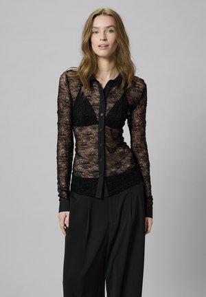 Schwarze Spitzenbluse mit durchgeknöpftem Vorderteil, durchsichtigen langen Ärmeln und festem Kragen. Dazu kombiniert mit hochgeschnittenen schwarzen Hosen.