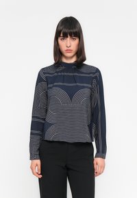Marinebluse mit geometrischen Linienmustern in Beige, mit hochgeschlossenem Kragen und langen Ärmeln, aus einem glatten Stoff gefertigt.