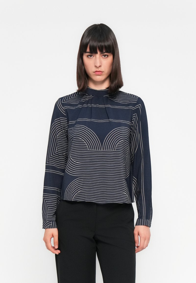 Marinebluse mit geometrischen Linienmustern in Beige, mit hochgeschlossenem Kragen und langen Ärmeln, aus einem glatten Stoff gefertigt.