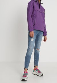 Lilla vandtæt jakke med halv lynlås foran, kengurulomme, kombineret med slidte blå jeans og flerfarvede sneakers.