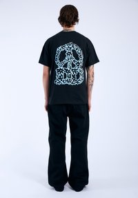 Camiseta negra con un gráfico de símbolo de paz en azul claro y un diseño floral en la parte trasera. Llevado con pantalones holgados negros.