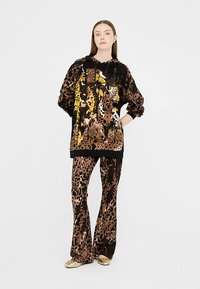 Oversizowa czarno-brązowa bluza z kapturem w zwierzęcy wzór z żółtymi akcentami, zestawiona z dopasowanymi rozszerzanymi spodniami w podobne zwierzęce wzory, noszona ze złotymi butami.