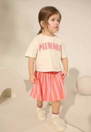 Lille pige med brunt hår iført en beige "MINNIE" trøje, pink og orange stribet nederdel, pink sokker og hvide sneakers, står i en neutral omgivelser.