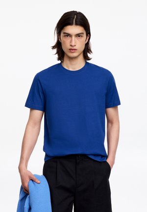 Joven con cabello oscuro de longitud media que lleva una camiseta azul y pantalones negros, sosteniendo un suéter azul doblado en su mano izquierda.