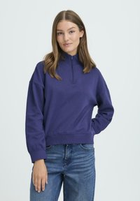 Vijolični pulover s polovičnim drsniki in dolgimi rokavi ter rebrastimi manšetami, v kombinaciji z modrimi kavbojkami. Mehek material in sproščen kroj.