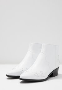 Botas de cuero blancas hasta el tobillo con puntas afiladas, costuras decorativas y tacones bajos negros sobre una superficie blanca con un fondo sencillo.