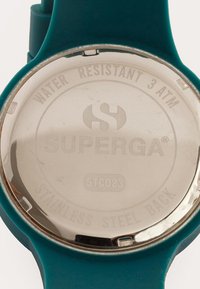 Türquoise kell, mille tagaküljel on roostevabast terasest graveeringud "SUPERGA," "WATER RESISTANT 3 ATM" ja mudelikood "STC023."