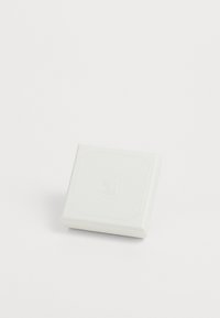 Valge tekstureeritud ruudukujuline kast, mille peal on väljaulatuv kaheksanurkne kuju ja reljeefne logo. Sujuvad servad ja minimalistik disain.