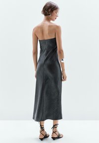 Schwarzes, trägerloses Maxikleid mit strukturiertem Finish, taillierter Form und einem Schlitz an der Seite. Getragen mit schwarzen Sandalen mit Riemchen und einem silbernen Armreif.