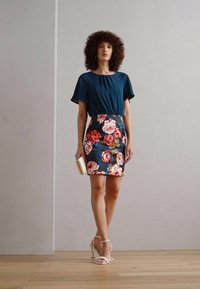 Türkise Bluse mit kurzen Ärmeln, kombiniert mit einem floralem Rock, der pinke und orange Rosen auf dunklem Hintergrund zeigt, ergänzt durch weiße Absätze und eine Clutch.