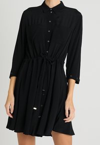 Schwarzes, langärmliges Hemdkleid mit Kragen und Taillengurt, in mittlerer Oberschenkelhöhe getragen.