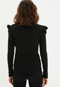 Czarny sweter prążkowany z falbankami na ramionach i długimi rękawami, z guzikami przy mankietach, o dopasowanym kroju dla uzyskania smukłej sylwetki.