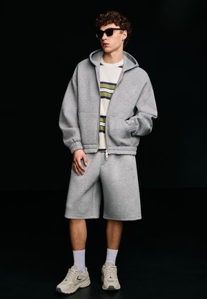 Jonge man met zwarte zonnebril, grijze zip-up hoodie, gestreept T-shirt, grijze wijde korte broek, witte sokken en beige sneakers tegen een zwarte achtergrond.