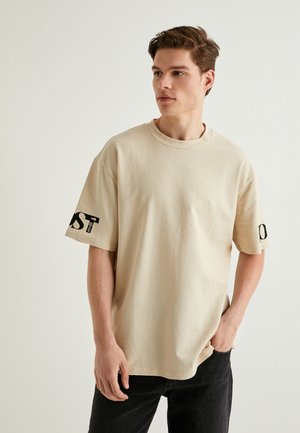 Beige kortærmet t-shirt i et tekstureret stof med rund hals, med kontrasterende sort skrift på venstre ærme.