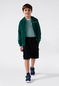 Χαμογελαστός αγόρι προχωράει φορώντας πράσινο φούτερ με φερμουάρ, ανοιχτό μπλε μπλουζάκι, μαύρες βερμούδες και μπλε αθλητικά παπούτσια σε λευκό φόντο.