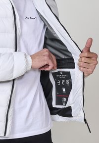 Giacca puffer bianca con fodera interna nera e tasca con zip che mostra le istruzioni per la cura e le icone. Materiale leggero e con texture.