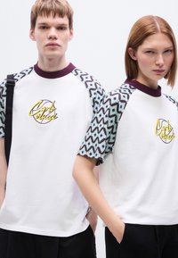 Μπλουζάκι unisex με λευκό σώμα, μανίκια σε μπλε με σχέδια και σκούρο καφέ, και μοβ ριμπ γιακά. Διαθέτει κίτρινο κεντημένο λογότυπο.