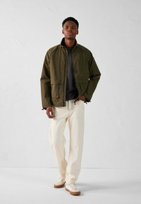 Veste légère vert olive avec un col montant, deux poches avant et des accents noirs, associée à un pantalon crème et des baskets beiges.
