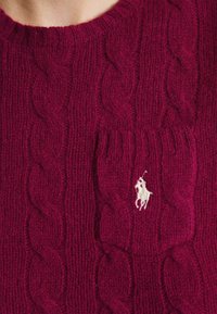 Stickad kabeltröja i djup vinröd färg, med rund halsringning och en liten vit broderad logga på den vänstra bröstfickan.