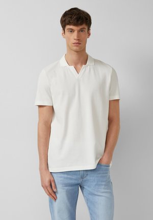 Junger Mann trägt ein weißes kurzärmeliges Poloshirt und hellblaue Jeans, steht mit einer Hand in der Tasche vor einem schlichten grauen Hintergrund.