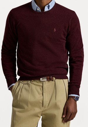 Man wearing a maroon sweater over a light blue collared shirt, beige pleated pants with a brown belt, and a silver watch on his left wrist.
