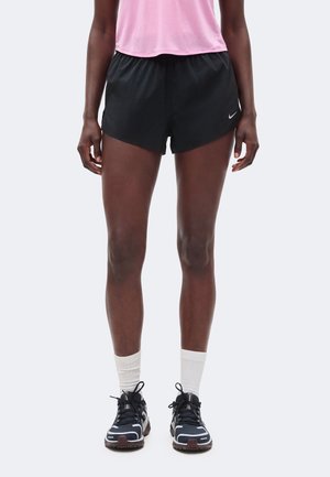 Osoba ubrana w czarne krótkie spodenki do biegania Nike, różowy top, białe skarpetki sportowe i czarne buty sportowe stojąca na jasnym, jednolitym tle.